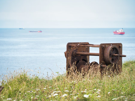 Rusted Industrial Winch on a Grassy Hill - A close-up, eye-level shot of a large, weathered, and rusted metal winch on a grassy clifftop, with the calm blue sea and cargo ships visible in the distance.の写真素材