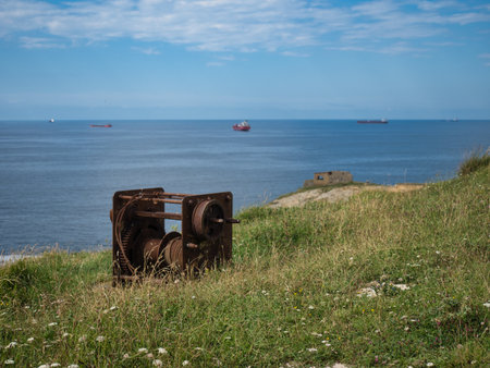 Rusted Industrial Winch on a Grassy Hill - A close-up, eye-level shot of a large, weathered, and rusted metal winch on a grassy clifftop, with the calm blue sea and cargo ships visible in the distance.の写真素材