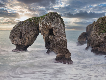 Natural Sea Arch in Rough Atlantic - Dramatic long exposure of a natural rock arch on the coast of Asturias, Spain, with turbulent white water and an overcast, moody sky.の写真素材