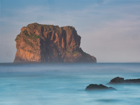 Sunset Light on a Coastal Island Stack - Majestic long exposure of a solitary sea stack or island bathed in warm sunset light, with a milky turquoise sea blurring the foreground in Asturias.の写真素材