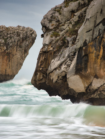 Hidden Beach and Jagged Sea Caves - Secluded beach between imposing limestone cliffs with sea caves and crashing waves on a dramatic, overcast day in Asturias, Spain.の写真素材