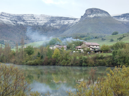 Snowy mountains over tranquil lake with farmhouses - A serene landscape with a lake reflecting farmhouses and a distant, snow-dusted mountain range under a winter sky.の写真素材