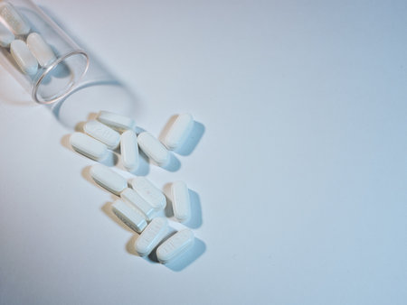 White Pills Spilled from Clear Bottle - Overhead shot of white oblong pills spilled from a clear plastic bottle on a light surface. Medical, pharmaceutical, and health concepts.の写真素材