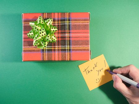 Thank You" Gift Box with Handwritten Note - Overhead view of a festive plaid gift box with a green bow and a hand writing a "Thank You!" note on a yellow sticky note, all on a green background. Perfect for holidays and appreciation.の写真素材