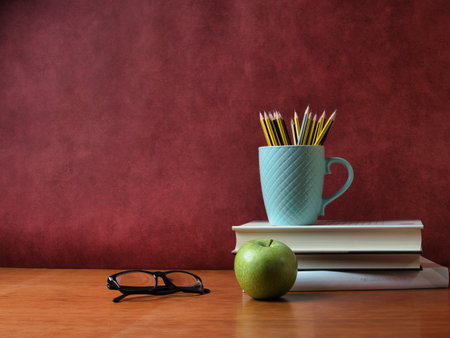 Vintage study desk setup with copy space - Glasses, apple, and pencils in a textured mug on a book stack and wooden desk. Educational concept with red background and space for text.の写真素材