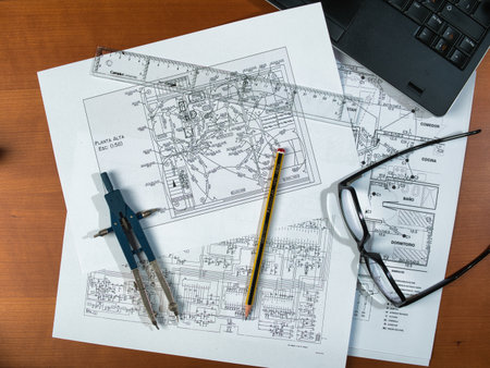 Overhead view of technical schematics and blueprints on a desk with glasses, a laptop, and a...