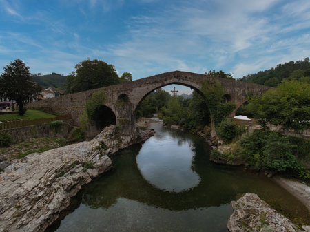 Historic Medieval Stone Bridge over River - Picturesque stone bridge with thick ivy crossing a calm, reflective river surrounded by lush green foliage and rocky banks.の写真素材