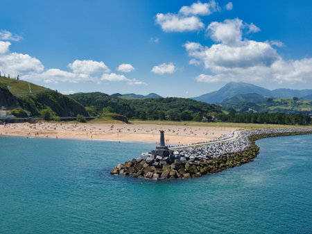 Coastal Breakwater and Beach on Sunny Day - Long, rocky breakwater and lighthouse leading into a port entrance next to a busy sandy beach and green rolling hills under a blue, cloudy sky.の写真素材