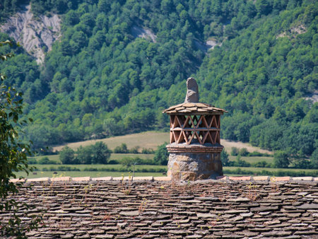 Mountain Village Stone Chimney on Tiled Roof - Traditional chimney on a slate tile roof with a forested mountain background on a sunny day.の写真素材