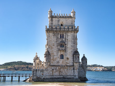Iconic BelÃ©m Tower on Tagus River, Lisbon - Full frontal view of the historic Torre de BelÃ©m fortress standing in the Tagus River, with a pier, the riverbank, and a clear blue sky.の写真素材