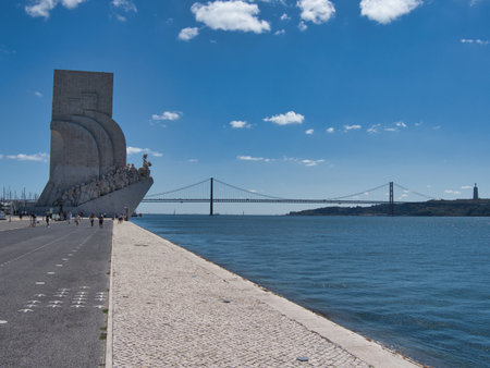 Discoveries Monument, Tagus River, Lisbon - The Padrão dos Descobrimentos monument on the Tagus riverfront, with the 25 de Abril Bridge and a clear blue sky in Lisbon, Portugal.の写真素材