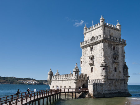 Iconic Belém Tower on Tagus River, Lisbon - Full frontal view of the historic Torre de Belém fortress standing in the Tagus River, with a pier, the riverbank, and a clear blue sky.の写真素材