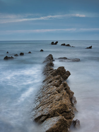 Striking Flysch Rock Leading Line Blue Hour Coast - Dramatic long exposure of a solitary flysch rock formation acting as a leading line towards the mountains under a cool blue hour sky.の写真素材