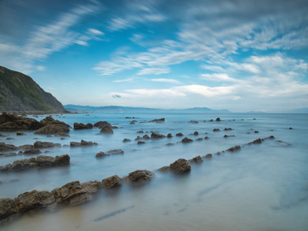 Serene Blue Sky Long Exposure Coastal Rocks - Beautiful long exposure shot of jagged flysch rock formations extending into a calm ocean under a bright, cloud-streaked blue sky.の写真素材