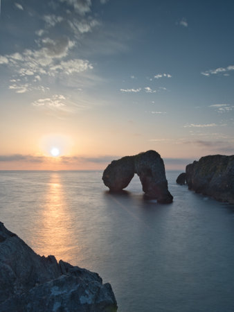 Sea Arch Silhouette at Blue Hour Twilight - Minimalist long exposure of a large sea arch and surrounding rock formations silhouetted against the still water and pastel pink and blue twilight sky.の写真素材