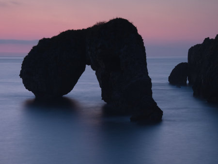 Sea Arch Silhouette at Blue Hour Twilight - Minimalist long exposure of a large sea arch and surrounding rock formations silhouetted against the still water and pastel pink and blue twilight sky.の写真素材