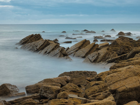 Jagged Flysch Rock Formations in Misty Ocean - Dramatic square-format long exposure of sharp flysch rock layers extending into the blurred, misty blue waters of the Atlantic ocean.の写真素材