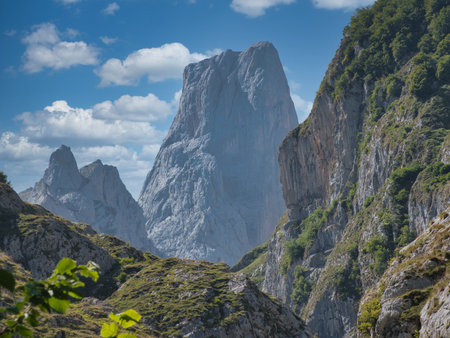 Majestic Naranjo de Bulnes Peak Under Blue Sky - A striking view of the towering limestone wall of Picu Urriellu against a vibrant blue sky with scattered white clouds, framed by rocky slopes.の写真素材