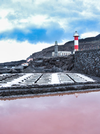 Lighthouse and Pink Salt Pans at Sunset - Striking coastal scene with the red and white lighthouse overlooking salt evaporation ponds with pink water reflection on volcanic terrain.の写真素材