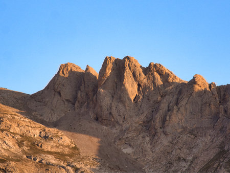 Golden hour on rugged mountain peaks - Dramatic shot of a rocky mountain range illuminated by warm golden hour light under a striking blue sky with scattered white clouds.の写真素材