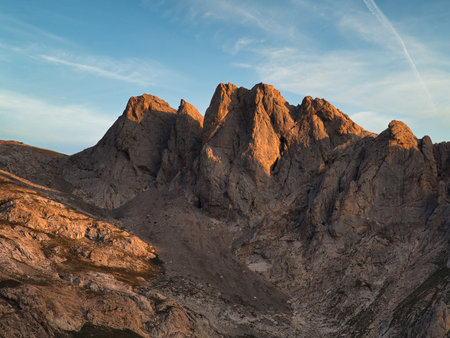 Golden hour on rugged mountain peaks - Dramatic shot of a rocky mountain range illuminated by warm golden hour light under a striking blue sky with scattered white clouds.の写真素材