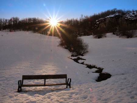 Sunburst over Snowy Winter Landscape with Bench - Lone wooden bench in a pristine snow-covered field with sun flares and starburst effect over a dark forest horizon.の写真素材