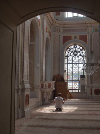 Worship and Sunlight in Mosque Interior - A man kneels in quiet prayer on the carpeted floor of a majestic mosque interior, illuminated by a dramatic shaft of sunlight through an ornate window.のeditorial素材