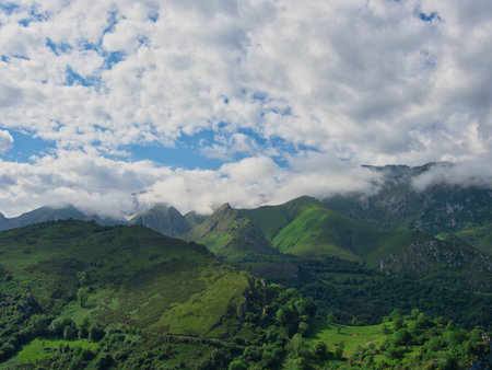 Lush green Picos de Europa rolling hills - Wide landscape view of a verdant mountain range with rolling green hills and dramatic clouds touching the peaks in Asturias, Spain.の写真素材