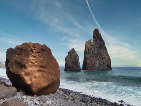 Giant Rock on Madeira's Pebbled Beach - Impressive low-angle view of the IlhÃ©us da Ribeira da Janela volcanic sea stacks in Madeira, seen from a rocky beach with a large foreground boulder and a contrail in the blue sky.の写真素材