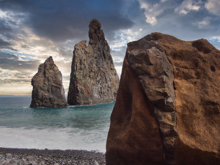 Giant Rock on Madeira's Pebbled Beach - Impressive low-angle view of the IlhÃ©us da Ribeira da Janela volcanic sea stacks in Madeira, seen from a rocky beach with a large foreground boulder and a contrail in the blue sky.の写真素材