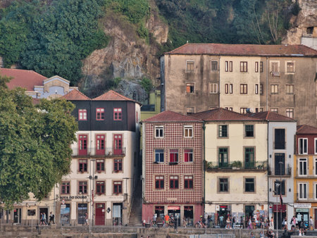 Historic Riverfront Buildings and Cliffside in Porto, Portugal - Row of traditional and colorful Ribeira district houses with red-tiled roofs nestled against a steep cliff face along the Douro River bank.の写真素材