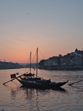 Rabelo Boats on Douro River at Dusk in Porto, Portugal - Silhouettes of Rabelo boats carrying barrels on the Douro River at sunset, with the historic Porto/Gaia cityscape dimly lit on the banks.の写真素材