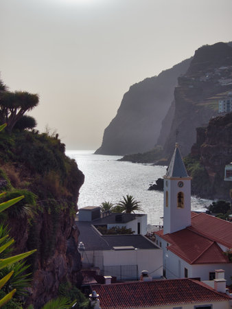 Coastal Church and Cliffs of Madeira, Portugal - Dramatic view of a whitewashed church spire and red rooftops framed by lush cliffs and the sparkling Atlantic Ocean in Madeira.の写真素材