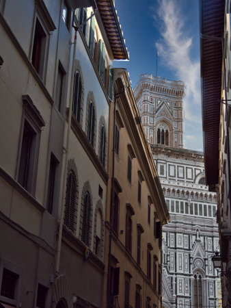 Duomo Facade and Giotto's Tower framed in Narrow Street - A unique view of the ornate marble facade and top of Giotto's Campanile, tightly framed between the shadowed, tall historic buildings of a Florence alley.の写真素材