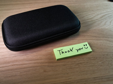 Thank You Note Next to Black Hard Case - Handwritten 'Thank You' note with a smiley face next to a black zippered hard case on a light wooden desk, conveying gratitude and appreciation.の写真素材