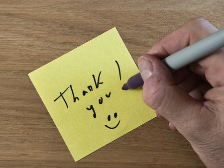 Thank You Note on Wooden Desk - Close-up of a hand writing 'Thank you!' with a marker on a yellow sticky note, placed on a textured wooden desk. Express gratitude and appreciation.の写真素材