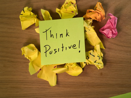 Think Positive! Note on Crumpled Paper - A vibrant sticky note displays 'Think Positive!' amidst a pile of crumpled yellow, orange, and pink notes on a wooden desk, symbolizing resilience.の写真素材