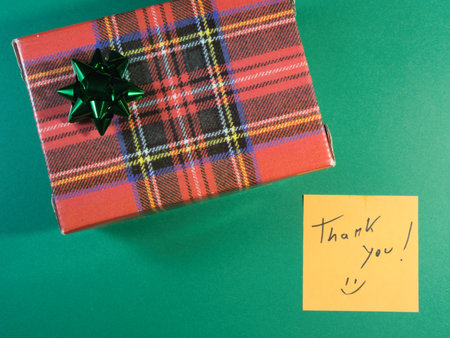Plaid Gift with Thank You Note - Top-down view of a red tartan gift box with a blue bow and a yellow 'Thank You!' sticky note on a vibrant green surface. Ideal for appreciation messages.の写真素材