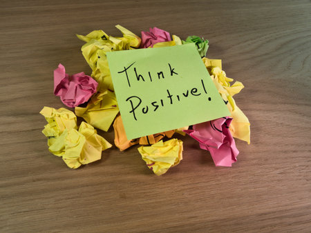 Think Positive Note on Crumpled Paper - An inspirational sticky note "Think Positive!" sits atop a pile of colorful crumpled papers on a wooden desk, symbolizing overcoming challenges.の写真素材