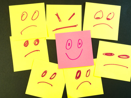 Happy Face Stands Out in Crowd of Sad - One pink sticky note with a happy face stands out from many yellow sticky notes displaying sad and angry expressions. Uniqueness.の写真素材