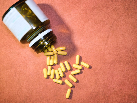 Yellow Capsules Spilling from Bottle - Vibrant yellow capsules spilling from an amber glass bottle onto a textured reddish-brown surface. Ideal for health, medicine, and wellness concepts.の写真素材