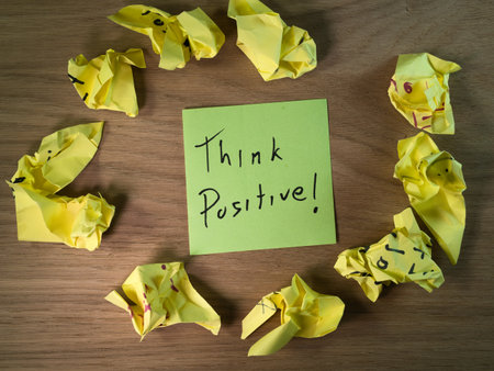 Think Positive! Green Note & Crumpled Paper - Green sticky note with 'Think Positive!' message surrounded by crumpled yellow papers on a wooden desk. Concept of positivity overcoming challenges.の写真素材