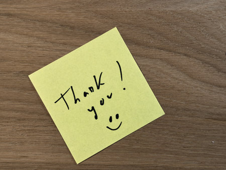 Handwritten Thank You on Yellow Sticky Note - Bright yellow sticky note with a handwritten 'Thank you!' message and smiley face on a warm, textured wooden desk background. Expressing gratitude.の写真素材
