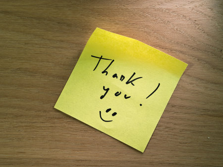 Handwritten Thank You on Yellow Sticky Note - Bright yellow sticky note with a handwritten 'Thank you!' message and smiley face on a warm, textured wooden desk background. Expressing gratitude.の写真素材