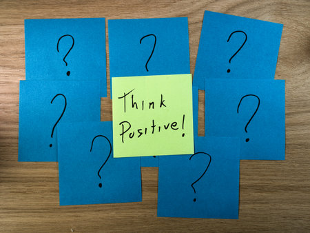 Think Positive! Amidst Questions - Handwritten 'Think Positive!' note centered among multiple question mark sticky notes on a wooden desk, representing hope, solutions, and challenges.の写真素材