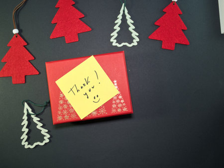 Thank You Gift Box with Christmas Decor - Red gift box with a yellow 'Thank you' note and festive Christmas tree ornaments on a dark background. Perfect for holiday gratitude messages.の写真素材