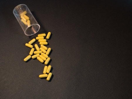 Medicine Bottle and Yellow Pills - An overhead shot of numerous bright yellow capsules, potentially prescription medication, scattered from an overturned clear plastic container on a solid black surface. Medical theme.の写真素材