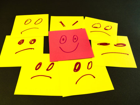 Happy Face Amidst Sad Faces - Red happy face sticky note stands out from numerous yellow sad and angry faces on a dark background, symbolizing positivity and uniqueness.の写真素材