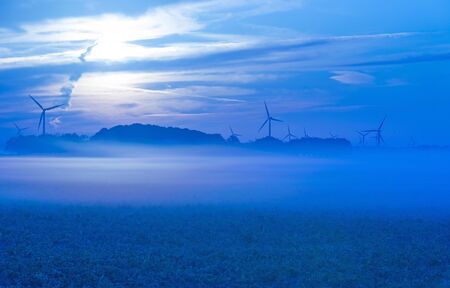 Sun shines on wind turbines in a foggy field at sunrise in summer