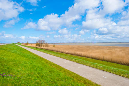 Road on a dike defying a stormy lake below a blue sky and white gray clouds in spring, Almere,...